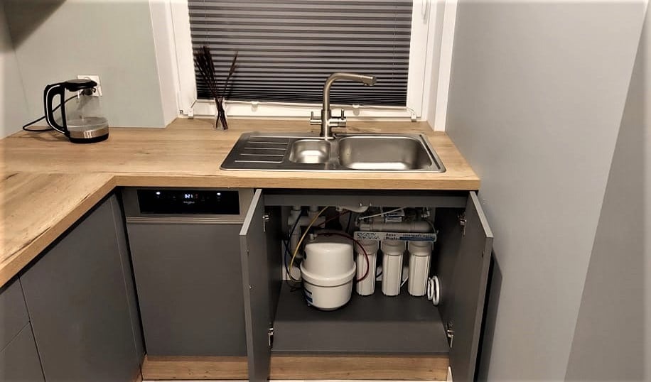 Filtr wody pod zlewem w szarej szafce kuchennej z drewnianym blatem, obok zmywarka. Widoczny kran i okno z żaluzjami. Filtr z kilkoma białymi cylindrami.