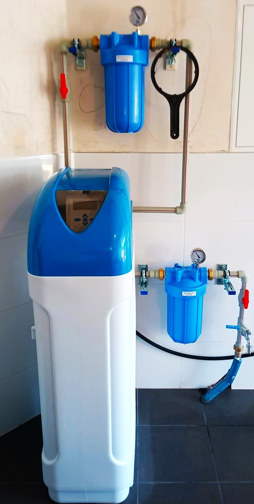 Kompletny system uzdatniania wody z filtrami i zmiękczaczem, zainstalowany na ścianie w pomieszczeniu z płytkami. Widoczne manometry i elementy instalacji.