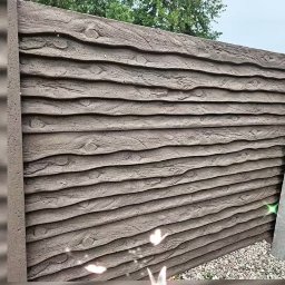 Firma Team Damian Podawczyk - Panelowy, betonowy płot imitujący drewno, z widoczną fakturą i detalami słojów, w tle zieleń drzew. Ułożony obok żwir.