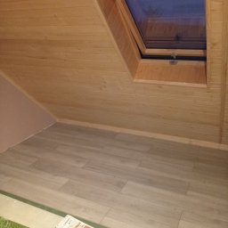 Firma Team Damian Podawczyk - Poddasze wykończone boazerią i panelami podłogowymi, z oknem dachowym. Widoczne gazety i ubrania na podłodze, sugerujące trwający remont lub wykończenie.