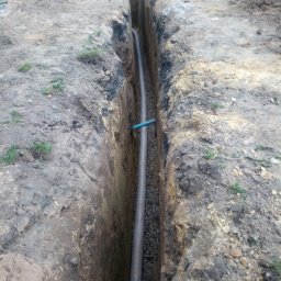 Firma Team Damian Podawczyk - Wykop z rurą drenażową wypełniony żwirem, widok z góry. Rura z niebieskim elementem łączeniowym. Na górze wykopu widoczne osoby i materiały budowlane.