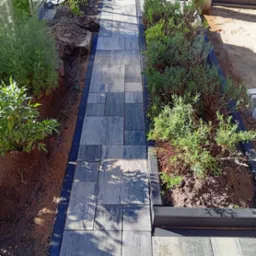 Ścieżka z szarej kostki brukowej o nieregularnym wzorze, obrzeża z czarnego tworzywa, zieleń po bokach. Widok z góry, oświetlenie słoneczne.