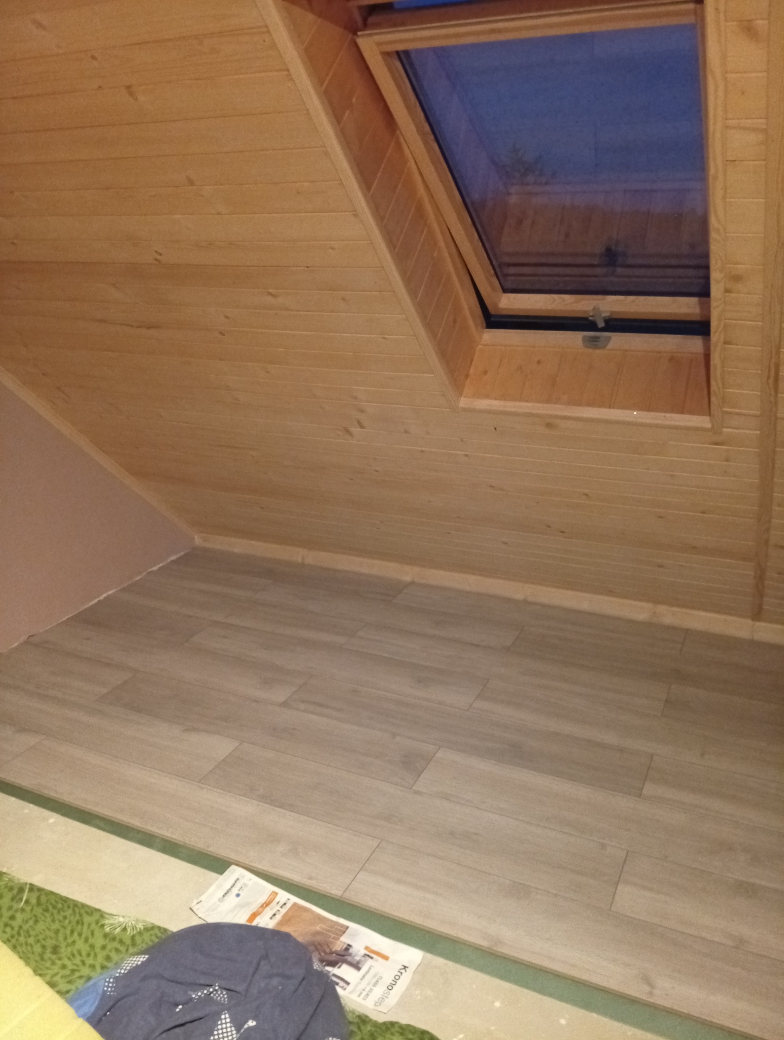 Poddasze wykończone boazerią i panelami podłogowymi, z oknem dachowym. Widoczne gazety i ubrania na podłodze, sugerujące trwający remont lub wykończenie.