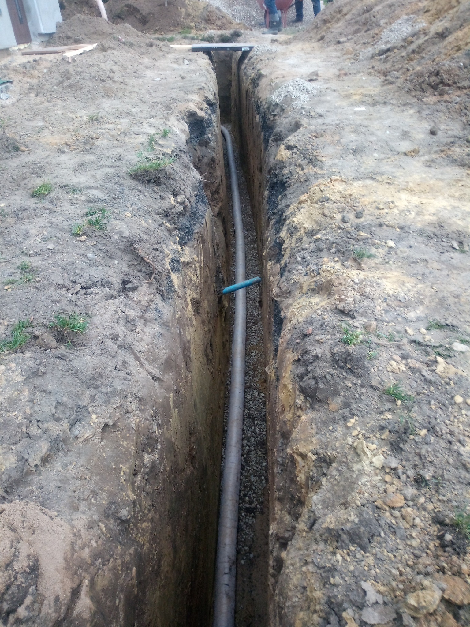 Wykop z rurą drenażową wypełniony żwirem, widok z góry. Rura z niebieskim elementem łączeniowym. Na górze wykopu widoczne osoby i materiały budowlane.