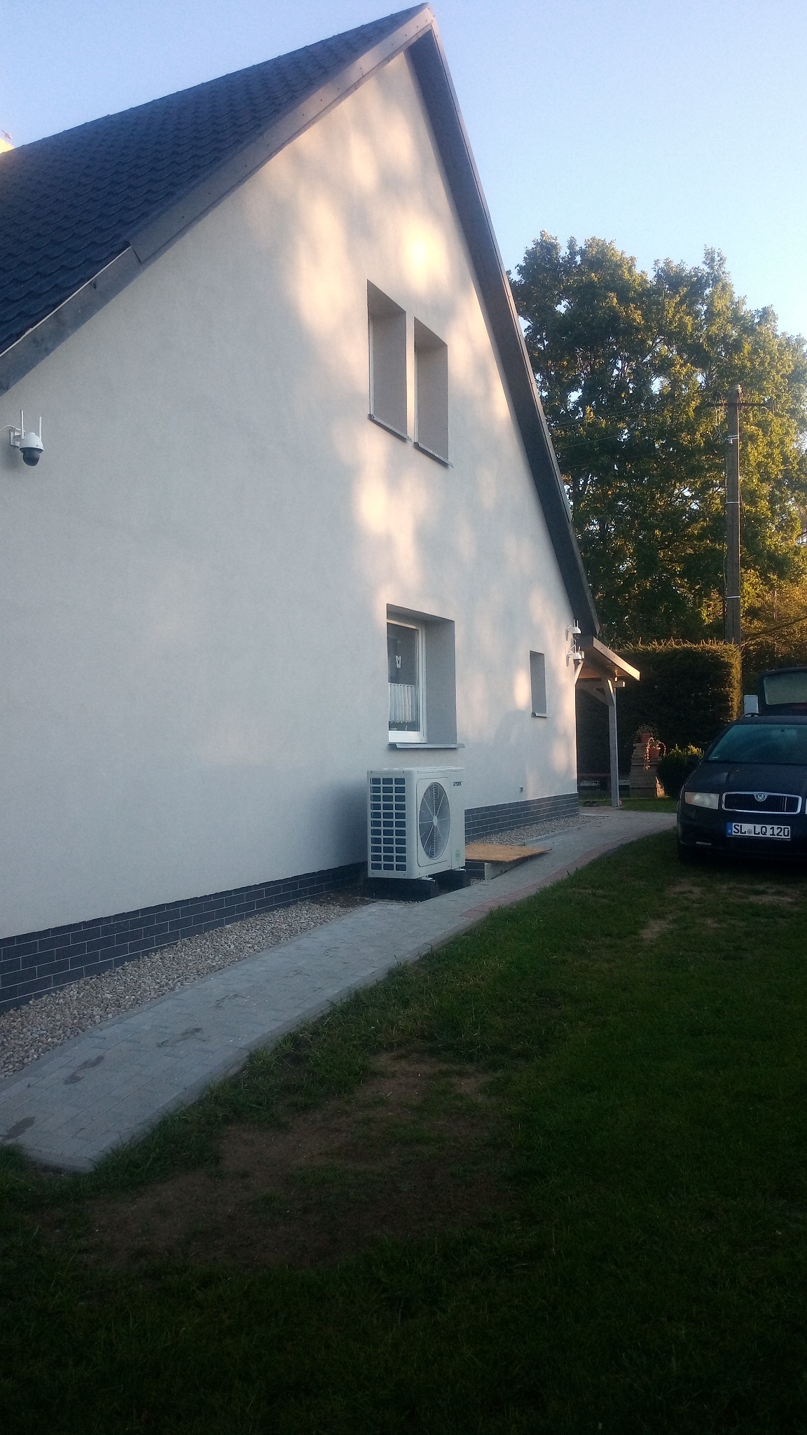 Elewacja domu z klimatyzatorem, widok z boku. Jasna fasada, szary dach, kostka brukowa i fragment trawnika. Z boku zaparkowany ciemny samochód osobowy.