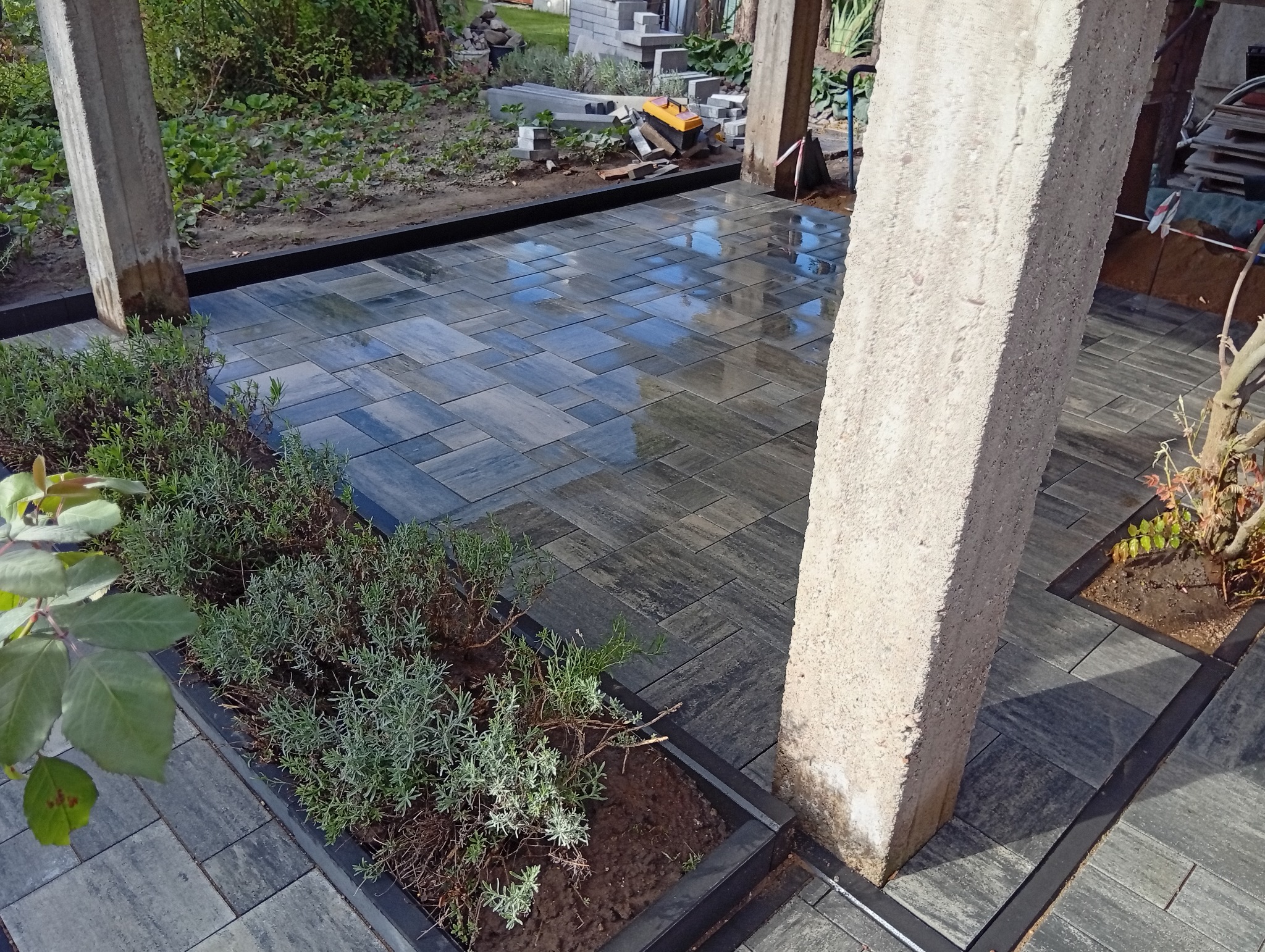 Ułożona, szara kostka brukowa z elementami zieleni w ogrodzie, widok z góry. Betonowe filary, czarne obrzeża, rośliny w grządkach.