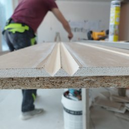 Wall Finish Łukasz Pacholski - Pracownik obrabia frezowaną płytę z widocznym przekrojem, w tle narzędzia i puszka z pianką. Płyta z wyfrezowanymi rowkami, przygotowywana do montażu.