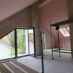 Wall Finish Łukasz Pacholski - Poddasze w trakcie prac wykończeniowych: ściany z płyt kartonowo-gipsowych, okna dachowe, rusztowania. Widok na zewnątrz przez duże okno.