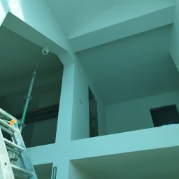 Wall Finish Łukasz Pacholski - Wnętrze w trakcie malowania, widoczne rusztowanie i świeżo pomalowane ściany na biało. Dwupoziomowe pomieszczenie z widocznymi przewodami elektrycznymi.
