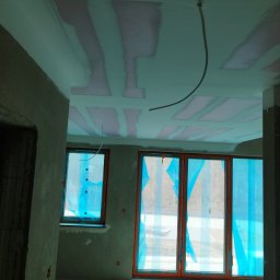 Wall Finish Łukasz Pacholski - Wnętrze w trakcie prac wykończeniowych: sufit z płyt karton-gips z widocznymi łączeniami, ściany przygotowane do malowania, okna zabezpieczone folią ochronną.