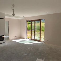 Wall Finish Łukasz Pacholski - Jasne wnętrze z drewnianymi drzwiami balkonowymi i widokiem na zieleń. Ściany w odcieniach beżu, surowa podłoga, widoczny kominek. Minimalistyczny wystrój, naturalne światło.