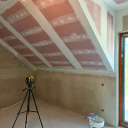 Wall Finish Łukasz Pacholski - Poddasze w trakcie prac wykończeniowych: widoczne płyty na skosach, szpachlowane łączenia, laser krzyżowy na statywie, surowa ściana i narzędzia w wiadrze.