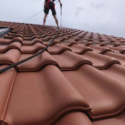Wall Finish Łukasz Pacholski - Czyszczenie dachówki ceramicznej z bliska. Widoczny pracownik w szelkach, używający lancy do mycia pod ciśnieniem. Dachówka w odcieniach brązu, mokra od wody.