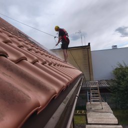 Wall Finish Łukasz Pacholski - Pracownik w kasku i uprzęży czyści dachówkę z brudu za pomocą myjki ciśnieniowej. Widoczny fragment dachu, drabina i elementy otoczenia.