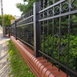 Eleganckie, czarne ogrodzenie z kutymi zdobieniami, osadzone na niskim murku z czerwonej cegły, otoczone zielenią. Widok z poziomu chodnika.