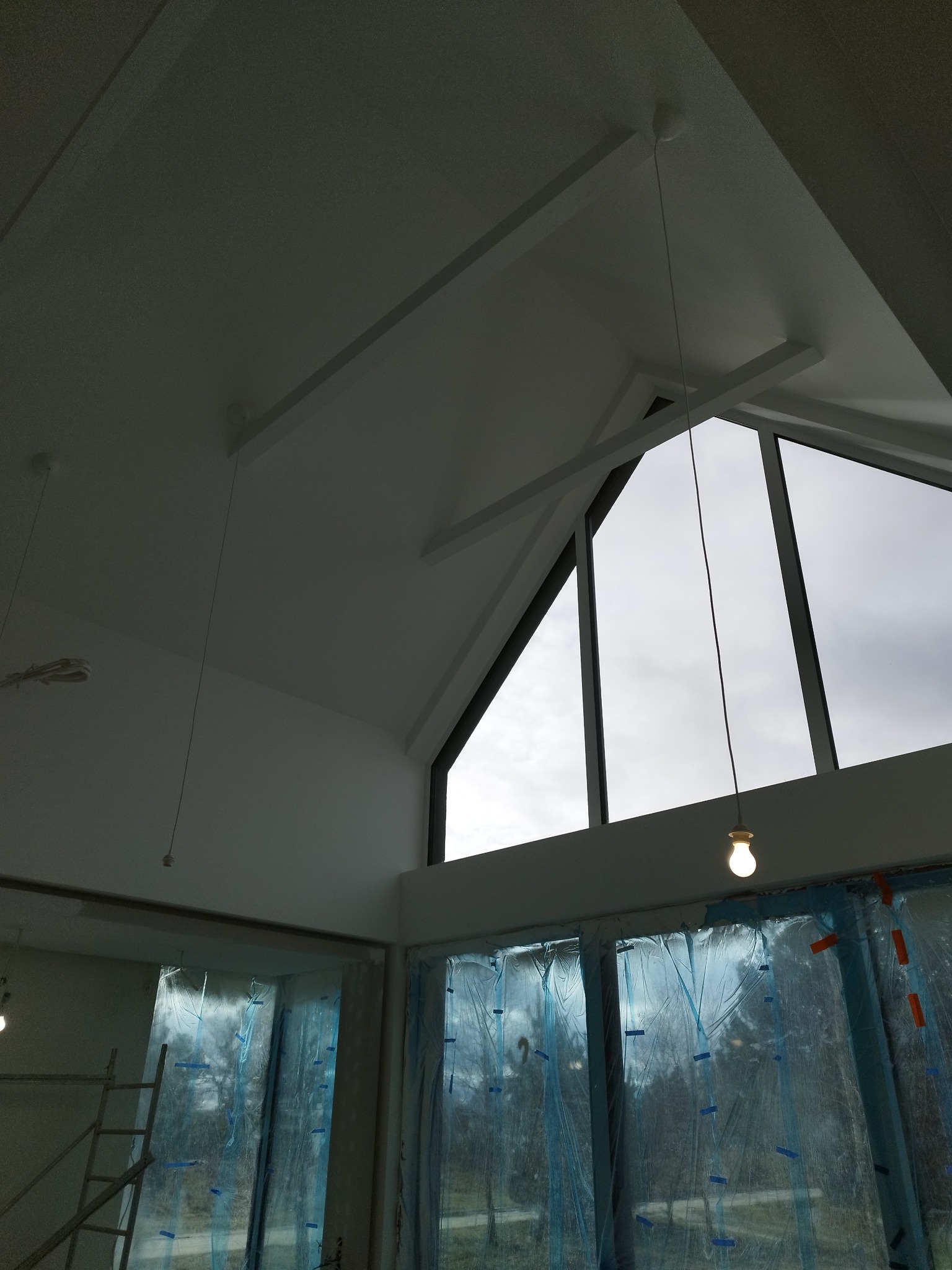 Jasne wnętrze z wysokim stropem i oknami w szczycie, zabezpieczonymi folią. Widoczne elementy konstrukcyjne i wiszące lampy. Etap prac wykończeniowych.