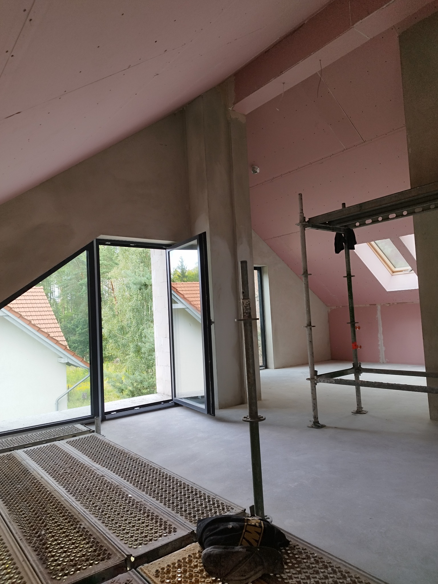 Poddasze w trakcie prac wykończeniowych: ściany z płyt kartonowo-gipsowych, okna dachowe, rusztowania. Widok na zewnątrz przez duże okno.