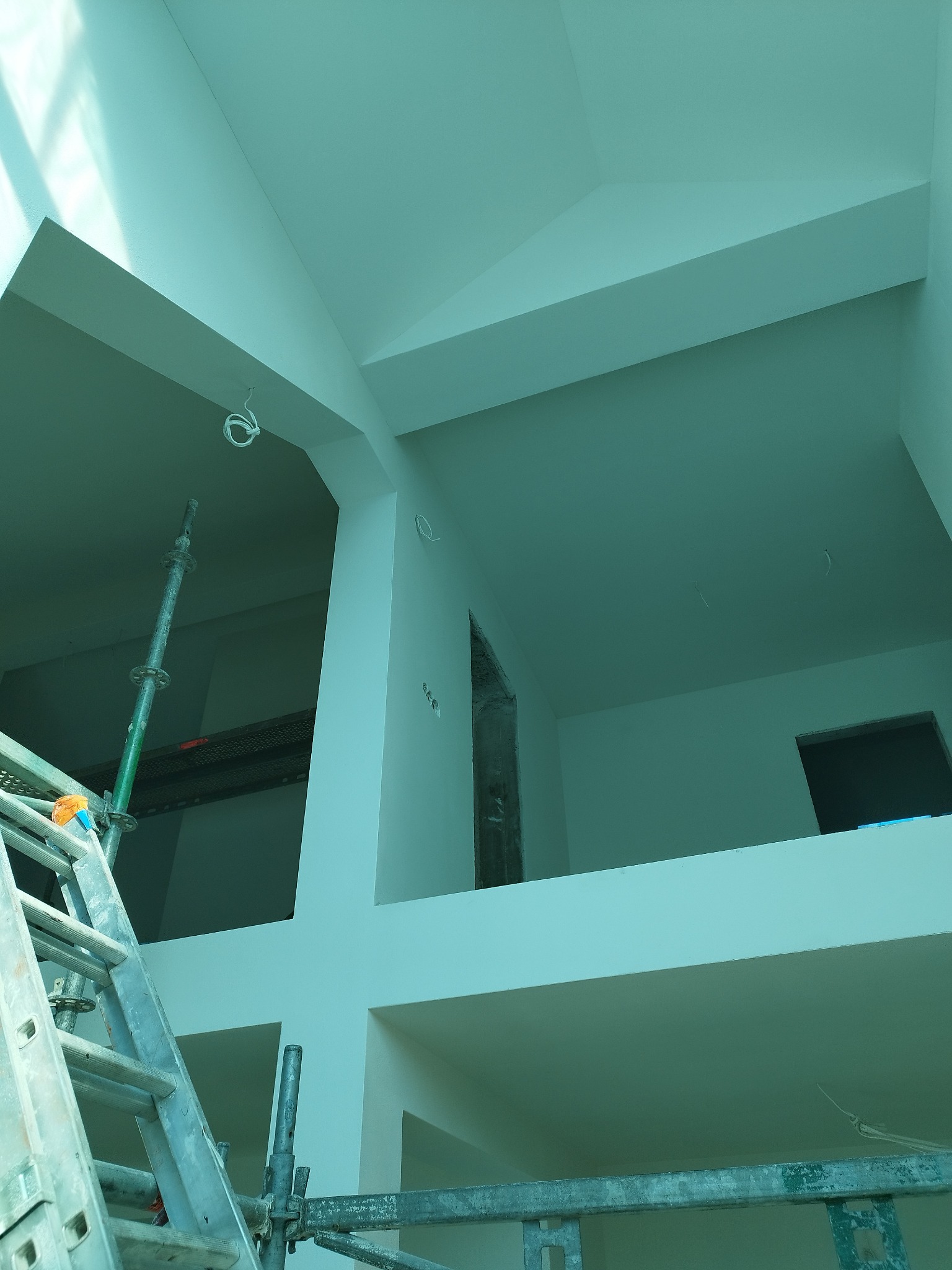 Wnętrze w trakcie malowania, widoczne rusztowanie i świeżo pomalowane ściany na biało. Dwupoziomowe pomieszczenie z widocznymi przewodami elektrycznymi.