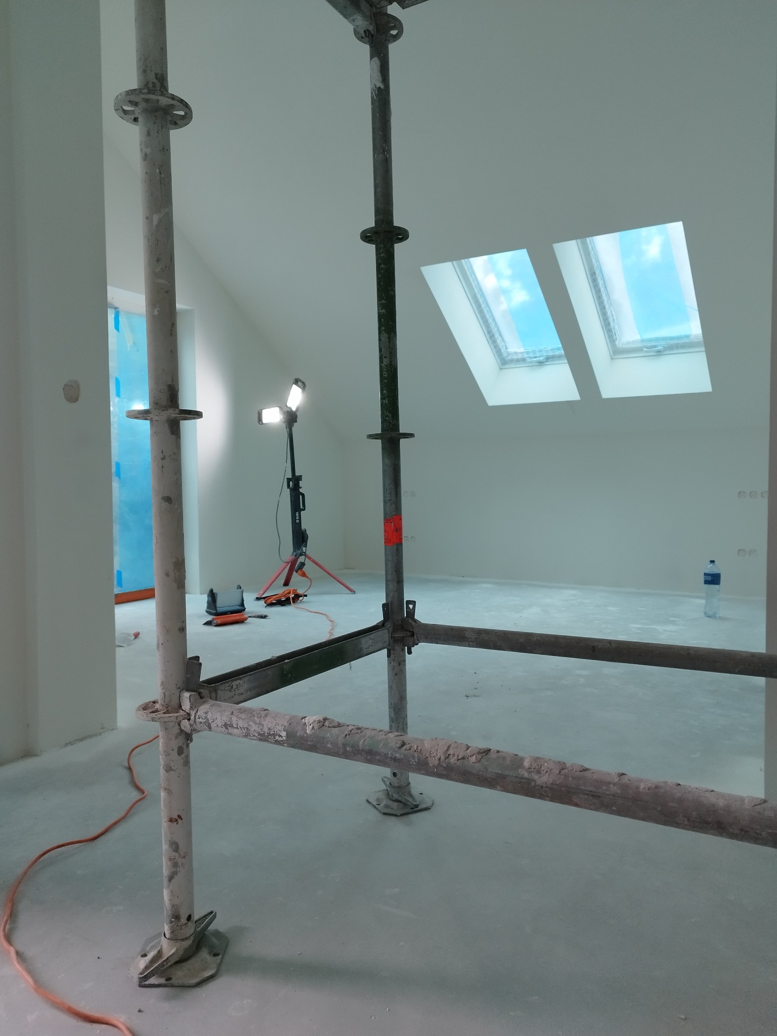 Poddasze w trakcie prac wykończeniowych. Widoczne rusztowanie, lampa budowlana i dwa okna dachowe w skosie. Ściany i podłoga przygotowane do dalszych prac.