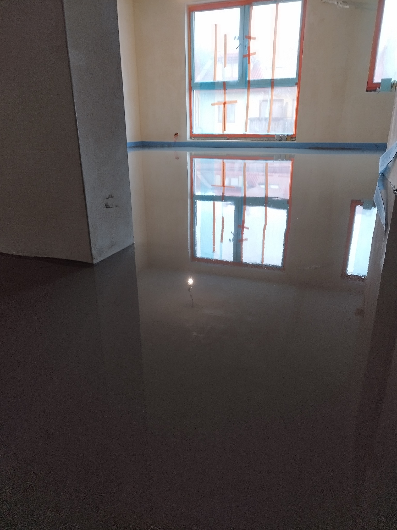 Wylana, gładka posadzka samopoziomująca w jasnym wnętrzu. Okna zabezpieczone folią malarską. Widoczny fragment ściany i kolumny. Błyszcząca powierzchnia odbija światło.