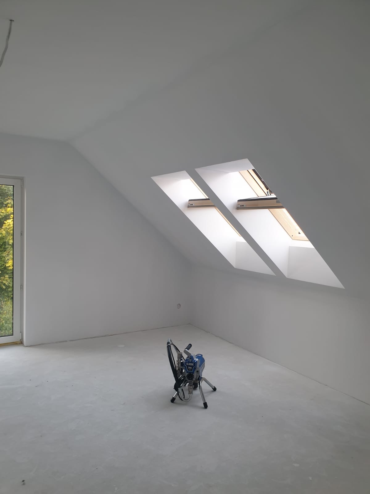 Poddasze po malowaniu na biało z oknami dachowymi. Na podłodze stoi agregat malarski. Widok z okna na zieleń. Świeżo wykończone wnętrze.