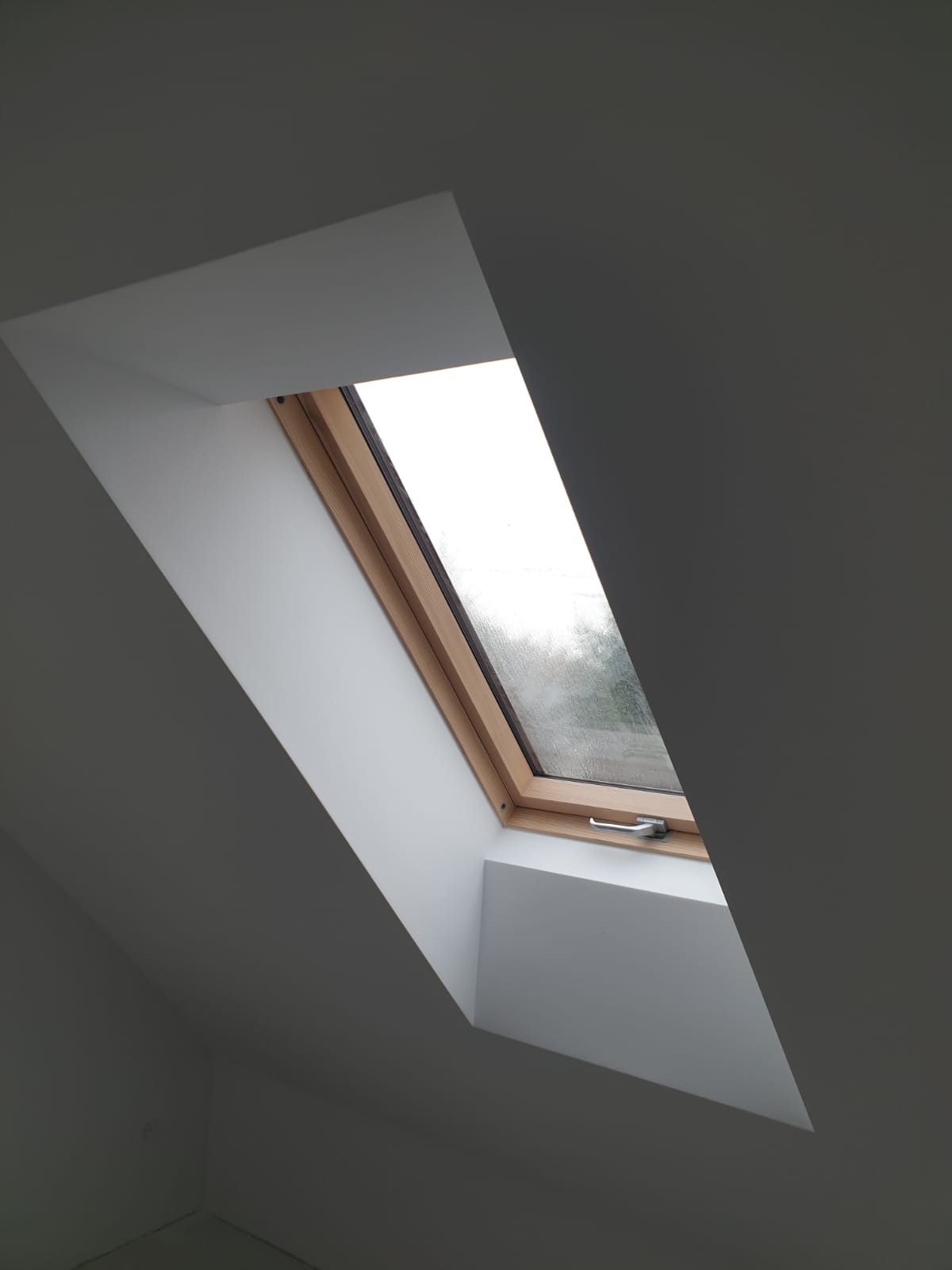 Okno dachowe z drewnianą ramą, osadzone w białej wnęce na szarym poddaszu. Widok z dołu, delikatne światło dzienne.