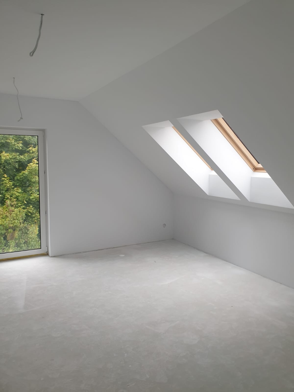 Poddasze po szpachlowaniu i malowaniu na biało, widoczne dwa okna dachowe i okno pionowe z widokiem na drzewa. Na suficie wystają kable.
