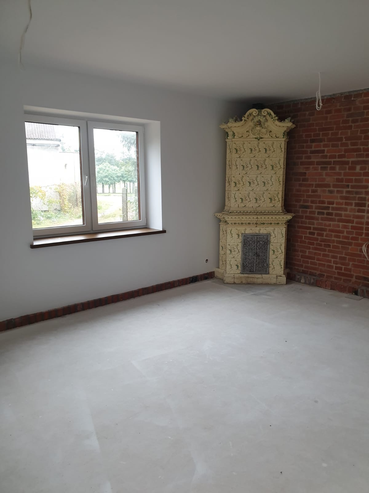 Pokój po remoncie: świeżo pomalowane ściany w odcieniach bieli, okno z drewnianym parapetem, ozdobny piec kaflowy i fragment ściany z czerwonej cegły.