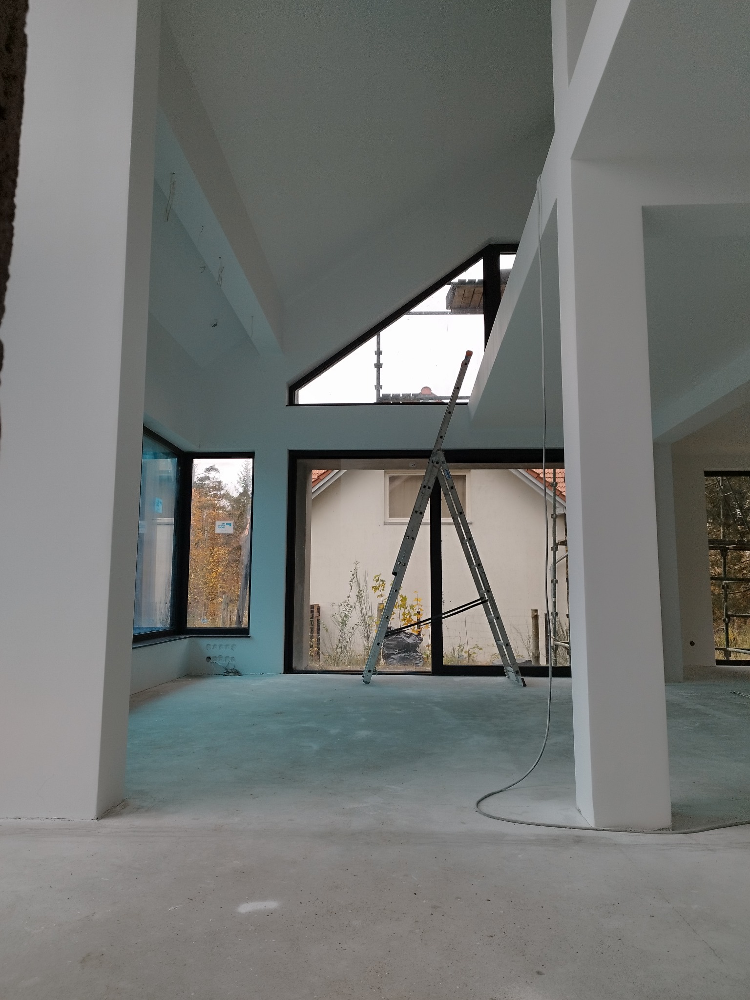 Wnętrze w trakcie wykańczania: białe ściany i kolumny, duże okna z widokiem na zewnątrz, drabina oparta o okno. Widoczne kable i surowa podłoga.