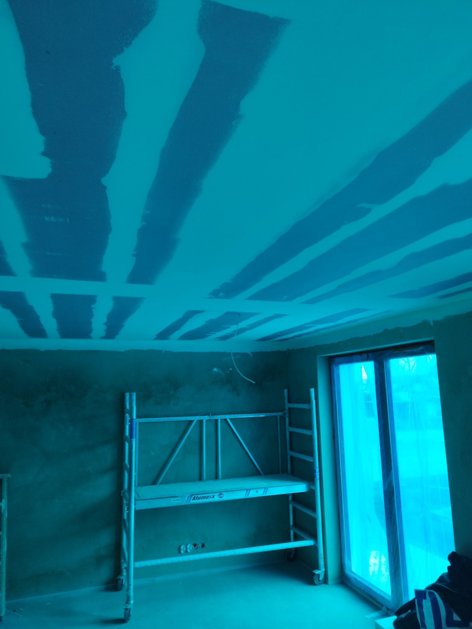 Wnętrze w trakcie prac wykończeniowych: sufit z widocznymi pasami szpachli, rusztowanie aluminiowe i okno zabezpieczone folią malarską. Całość w chłodnej tonacji.