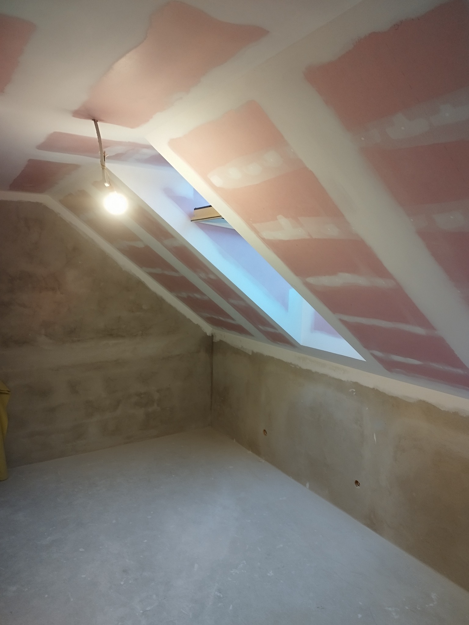 Poddasze ze skośnym oknem dachowym w trakcie prac wykończeniowych. Widoczne płyty kartonowo-gipsowe na suficie i przygotowane ściany, oświetlone wiszącą żarówką.