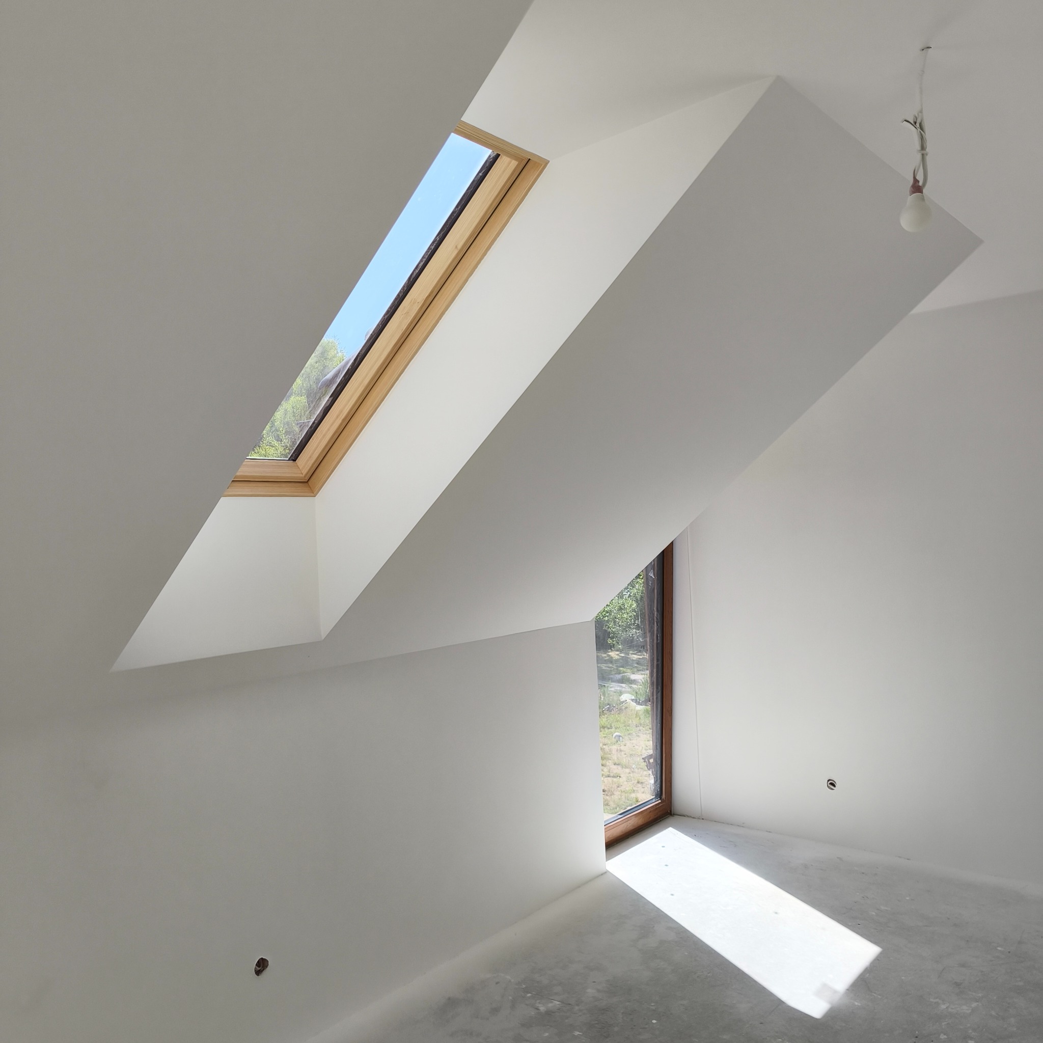 Jasne poddasze z oknem dachowym i pionowym. Świeżo pomalowane ściany na biało, surowa podłoga. Widoczne punkty elektryczne. Minimalizm i prostota.