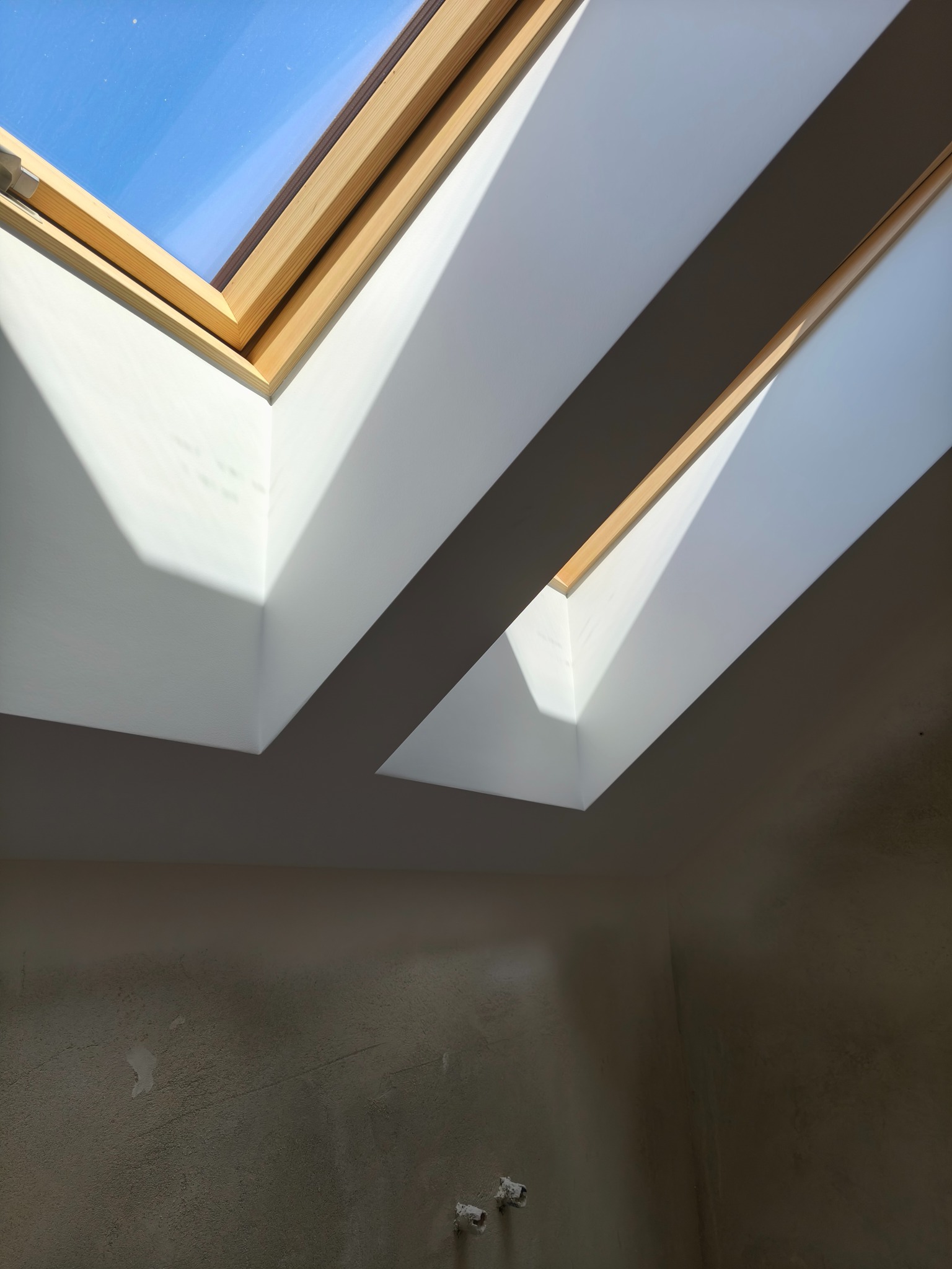 Wnętrze z oknami połaciowymi w jasnej obudowie i pomalowanymi na biało wnękami. Ściana w dolnej części surowa, przygotowana do wykończenia.