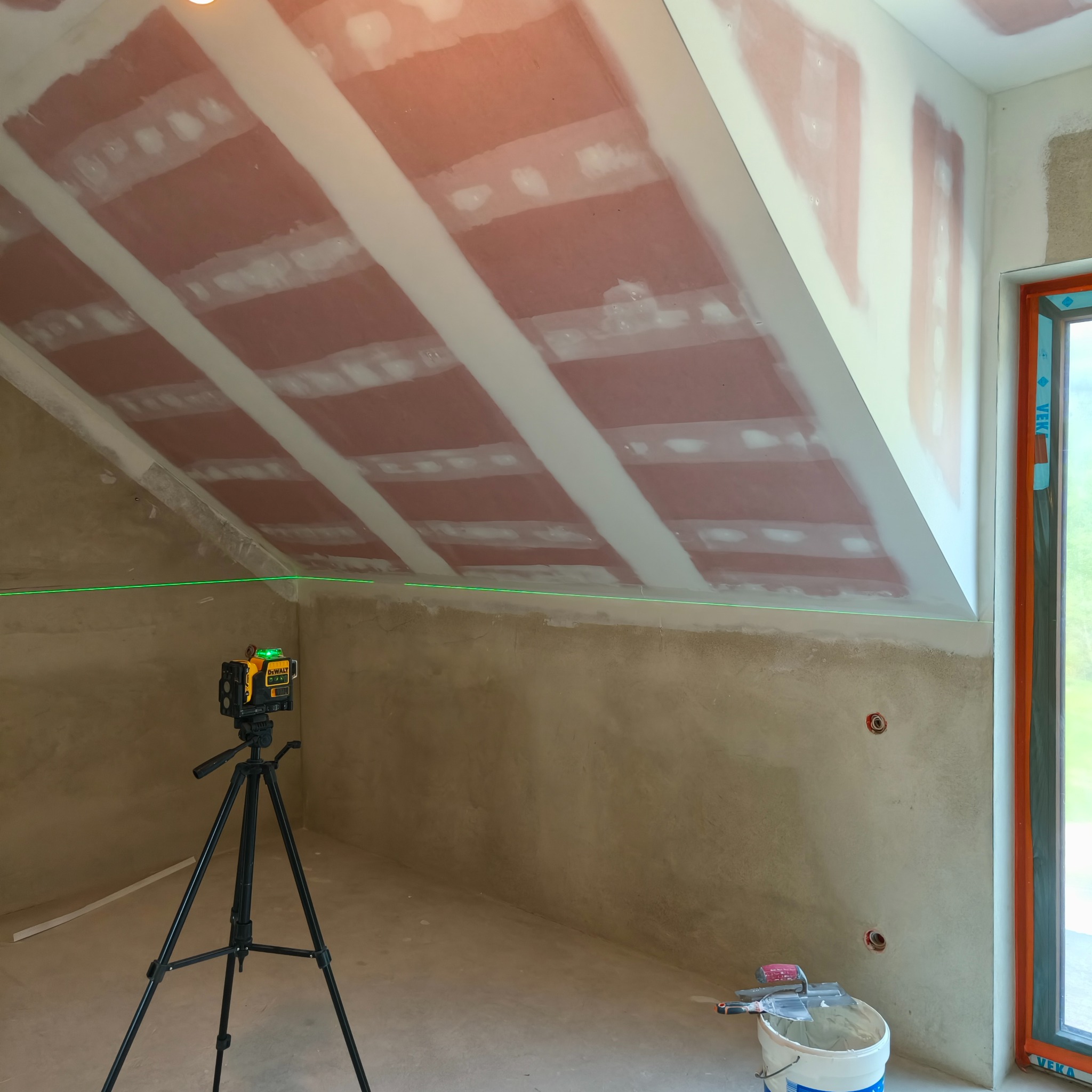 Poddasze w trakcie prac wykończeniowych: widoczne płyty na skosach, szpachlowane łączenia, laser krzyżowy na statywie, surowa ściana i narzędzia w wiadrze.