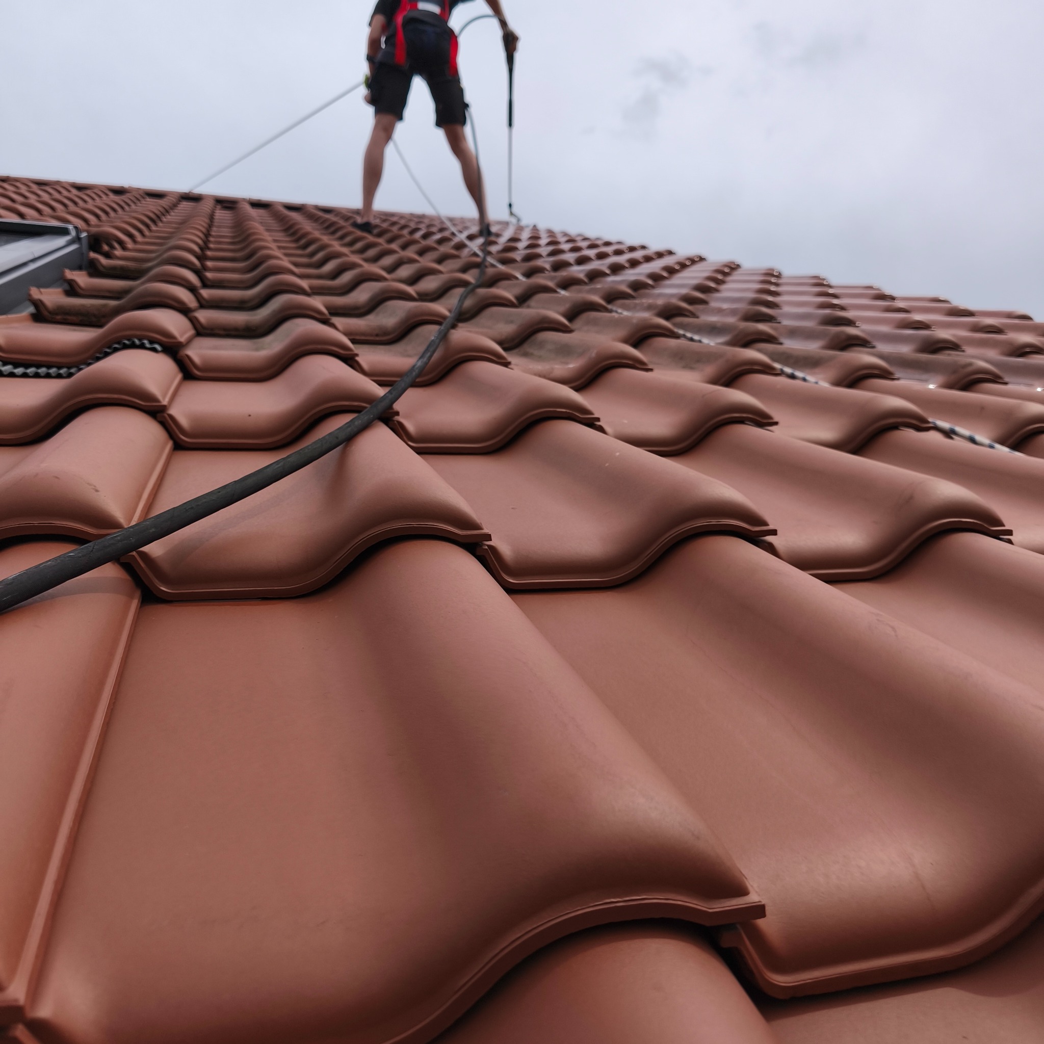 Czyszczenie dachówki ceramicznej z bliska. Widoczny pracownik w szelkach, używający lancy do mycia pod ciśnieniem. Dachówka w odcieniach brązu, mokra od wody.