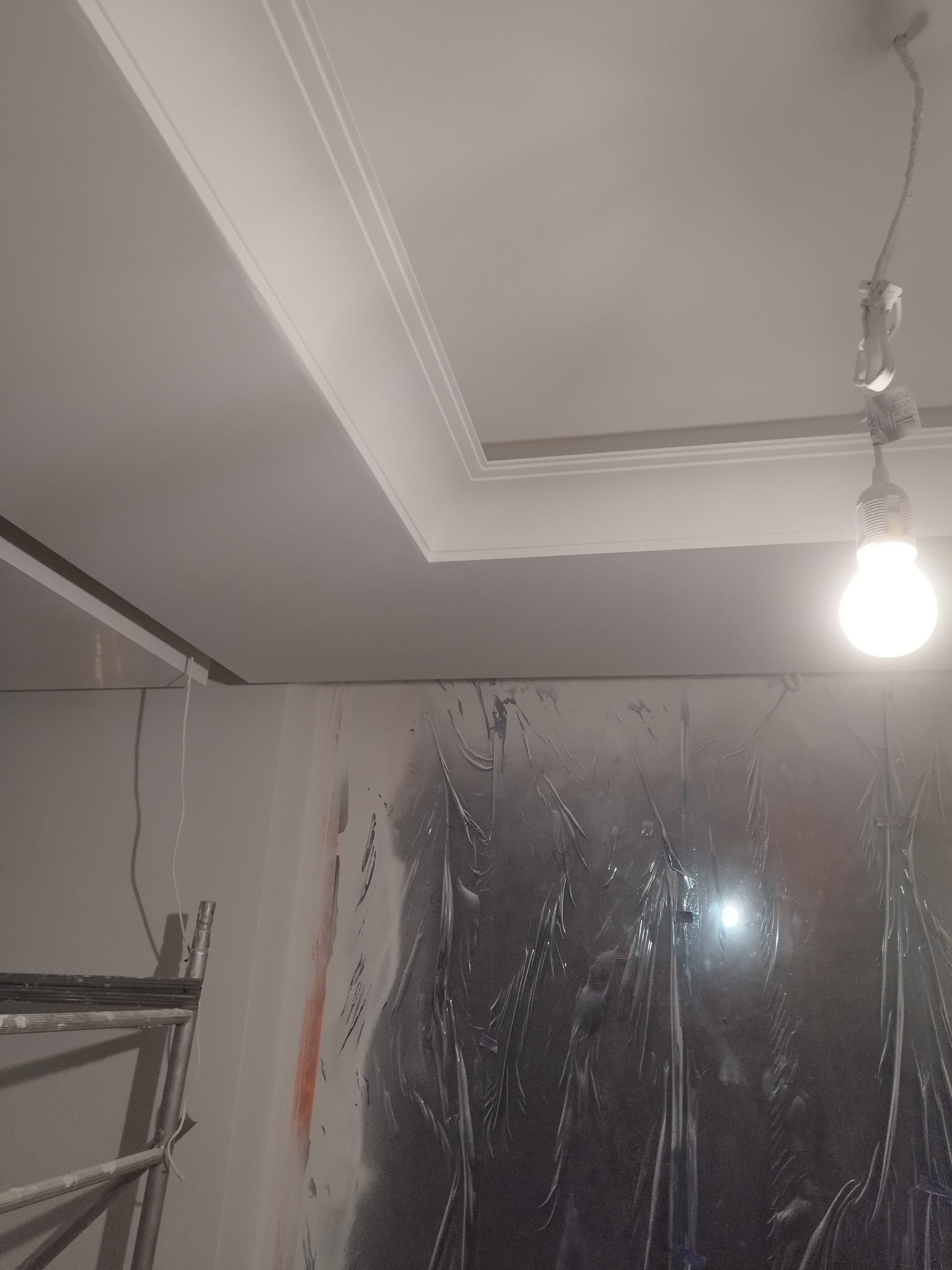 Wykończony sufit z dekoracyjną listwą, zabezpieczona folią ściana, drabina i żarówka na kablu. Ujęcie z dołu ukazuje detale wykończenia.