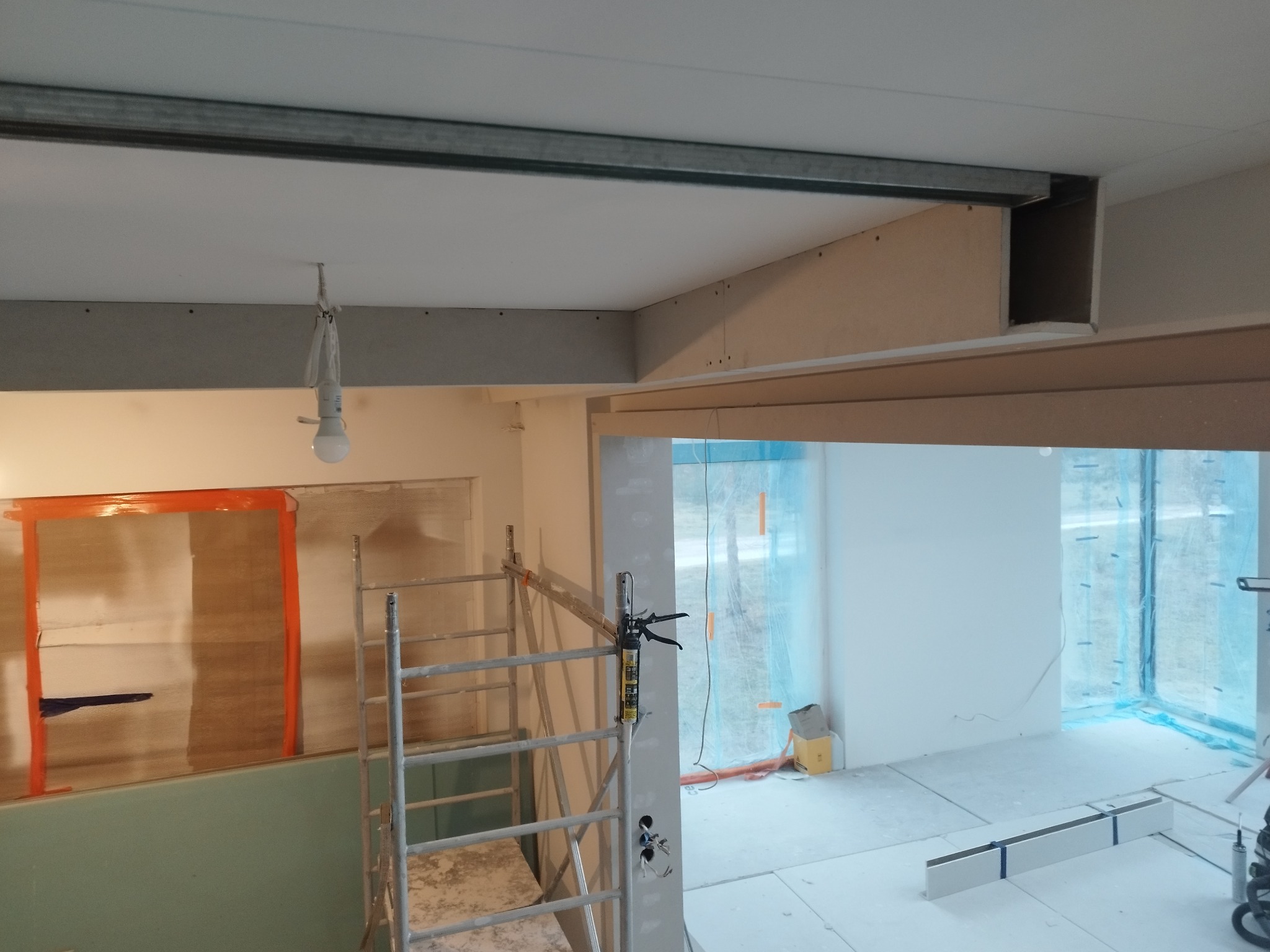 Wnętrze w trakcie prac wykończeniowych: widoczne stelaże sufitowe, częściowo zamontowane płyty, rusztowanie i narzędzia. Świecąca żarówka na kablu.