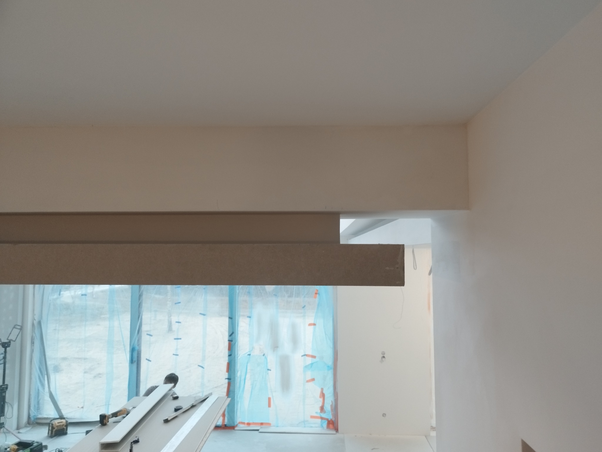 Wnętrze w trakcie wykańczania: widoczne elementy konstrukcji sufitu z płyt, zabezpieczone okna folią i narzędzia na stole, sugerujące prace wykończeniowe.
