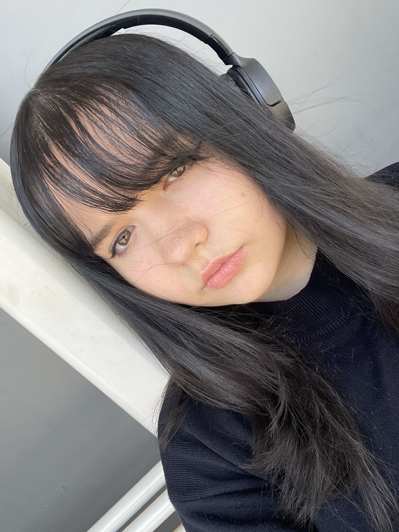 Portret kobiety z długimi, ciemnymi włosami i słuchawkami na głowie, opierającej się o białą balustradę, z delikatnym makijażem i naturalnym oświetleniem.