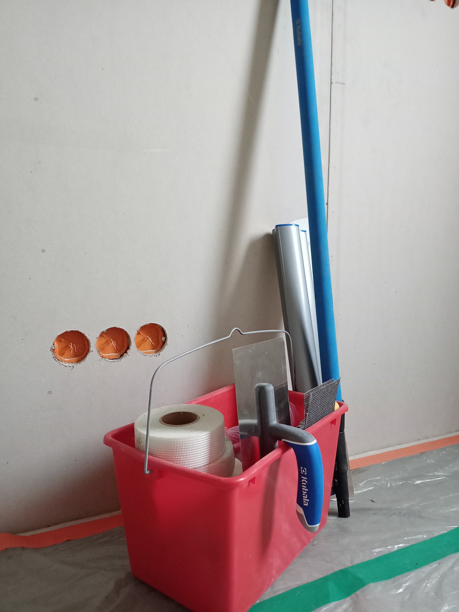 Czerwone wiadro z narzędziami malarskimi (szpachelka, taśma) stoi na folii ochronnej przy ścianie z trzema otworami na gniazdka. Widoczne akcesoria marki Kubala.