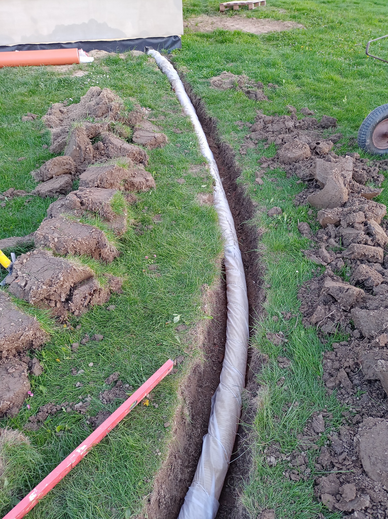 Drenaż liniowy w wykopie na trawniku. Rura drenażowa owinięta geowłókniną. Widoczne narzędzia i wykopana ziemia. Etap prac ziemnych przed zasypaniem.