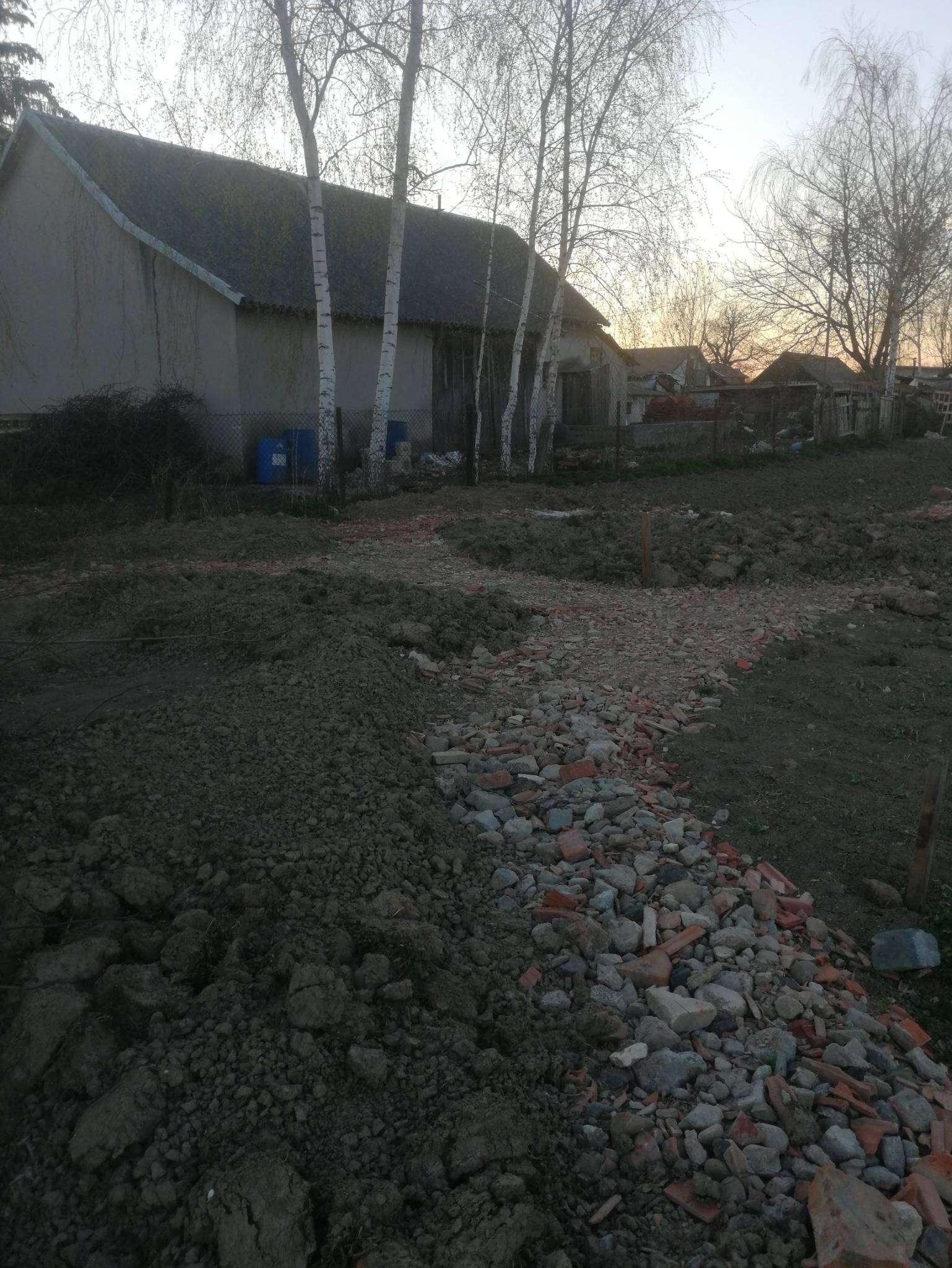 Przygotowanie terenu pod ścieżkę w ogrodzie. Widoczne gruz, kamienie i ziemia. W tle budynek gospodarczy i brzozy.