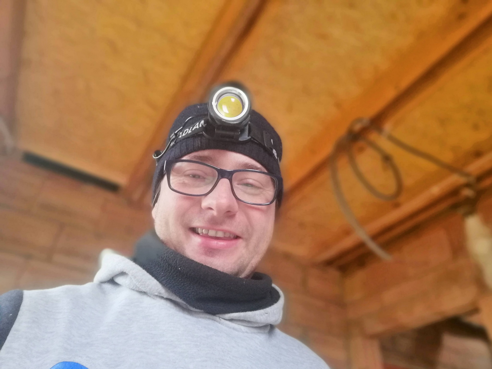 Uśmiechnięty mężczyzna w czapce i okularach z lampą czołową, prawdopodobnie podczas pracy wewnątrz budynku w stanie surowym.