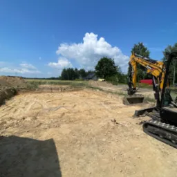 Żółta mini koparka SANY na wykopie pod fundamenty, w tle błękitne niebo z chmurami, teren budowy. Widoczny wykop w ziemi i sterta ziemi obok.