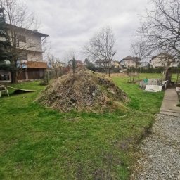 Usługi mini koparką Pszczyna - Kupa ziemi i roślin w ogrodzie, częściowo na trawie, z widocznymi budynkami w tle. Pochmurne niebo. Kable w lewym dolnym rogu kadru.