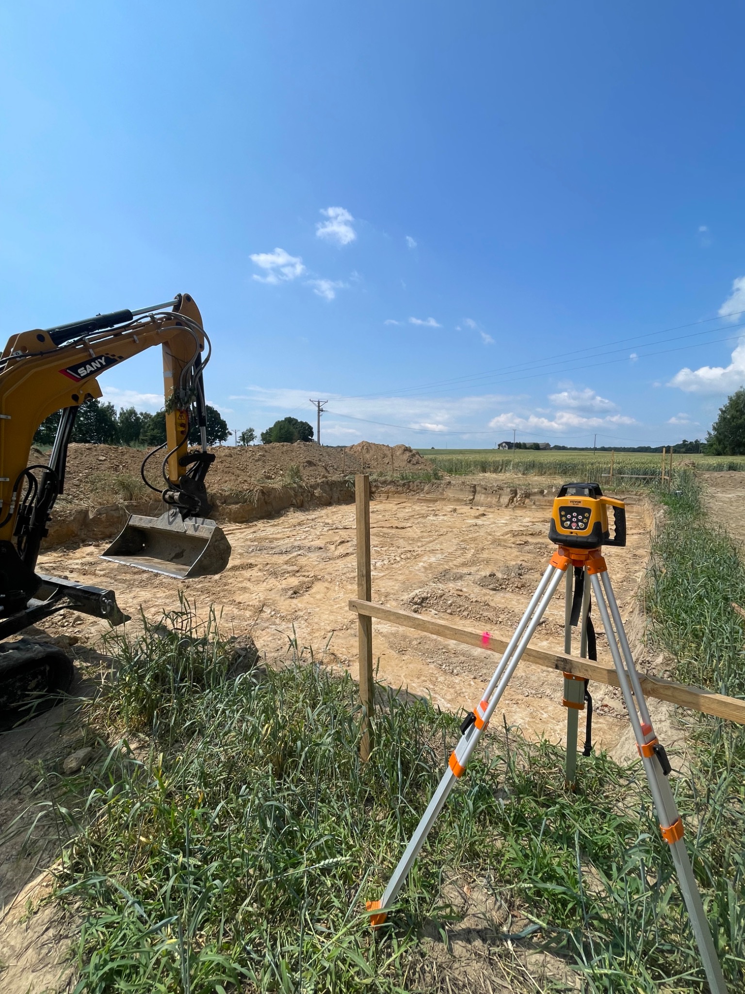 Żółta koparka SANY i niwelator laserowy na statywie obok wykopu pod fundamenty, otoczonego drewnianym ogrodzeniem, w słoneczny dzień.