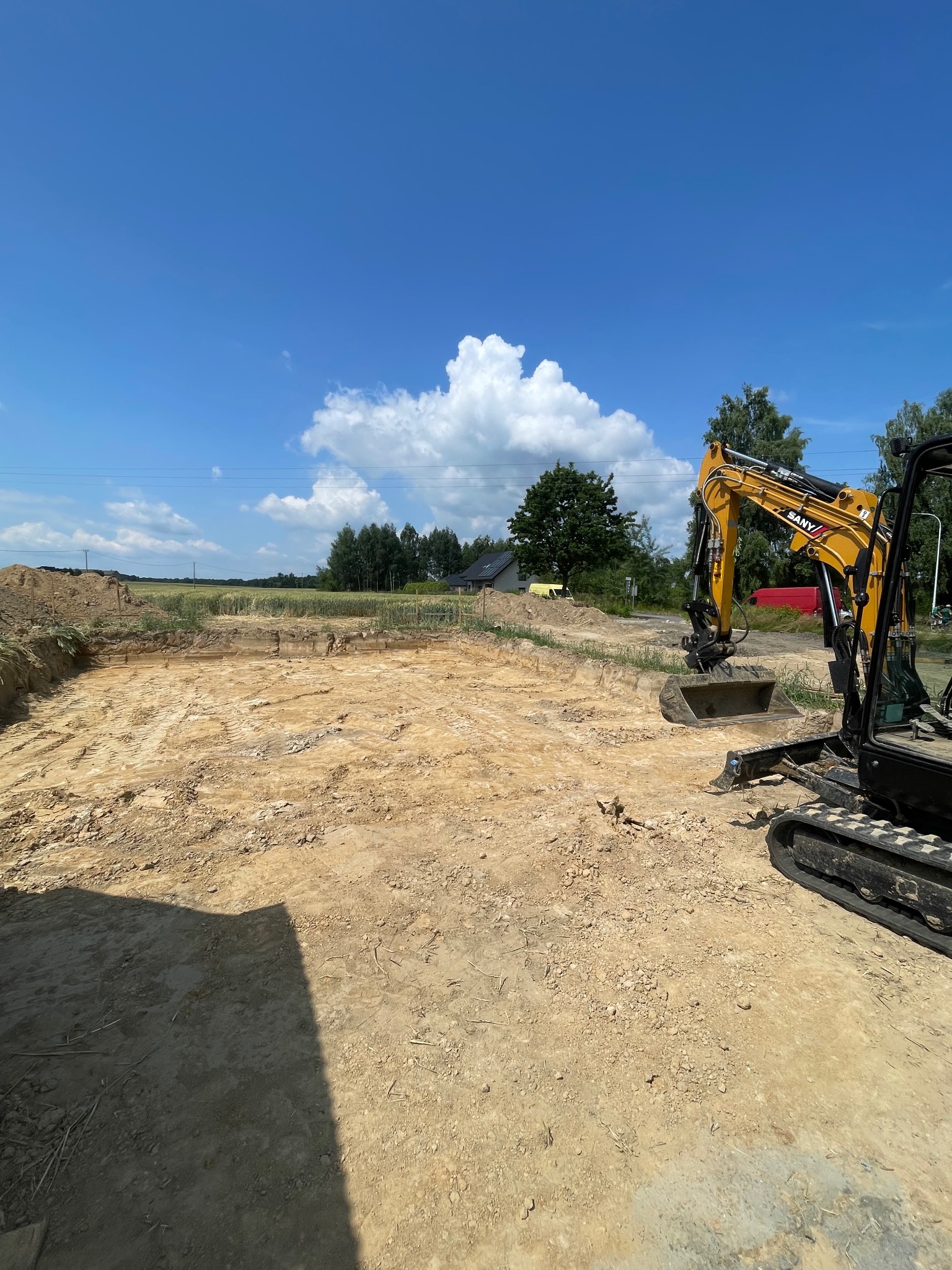 Żółta mini koparka SANY na wykopie pod fundamenty, w tle błękitne niebo z chmurami, teren budowy. Widoczny wykop w ziemi i sterta ziemi obok.