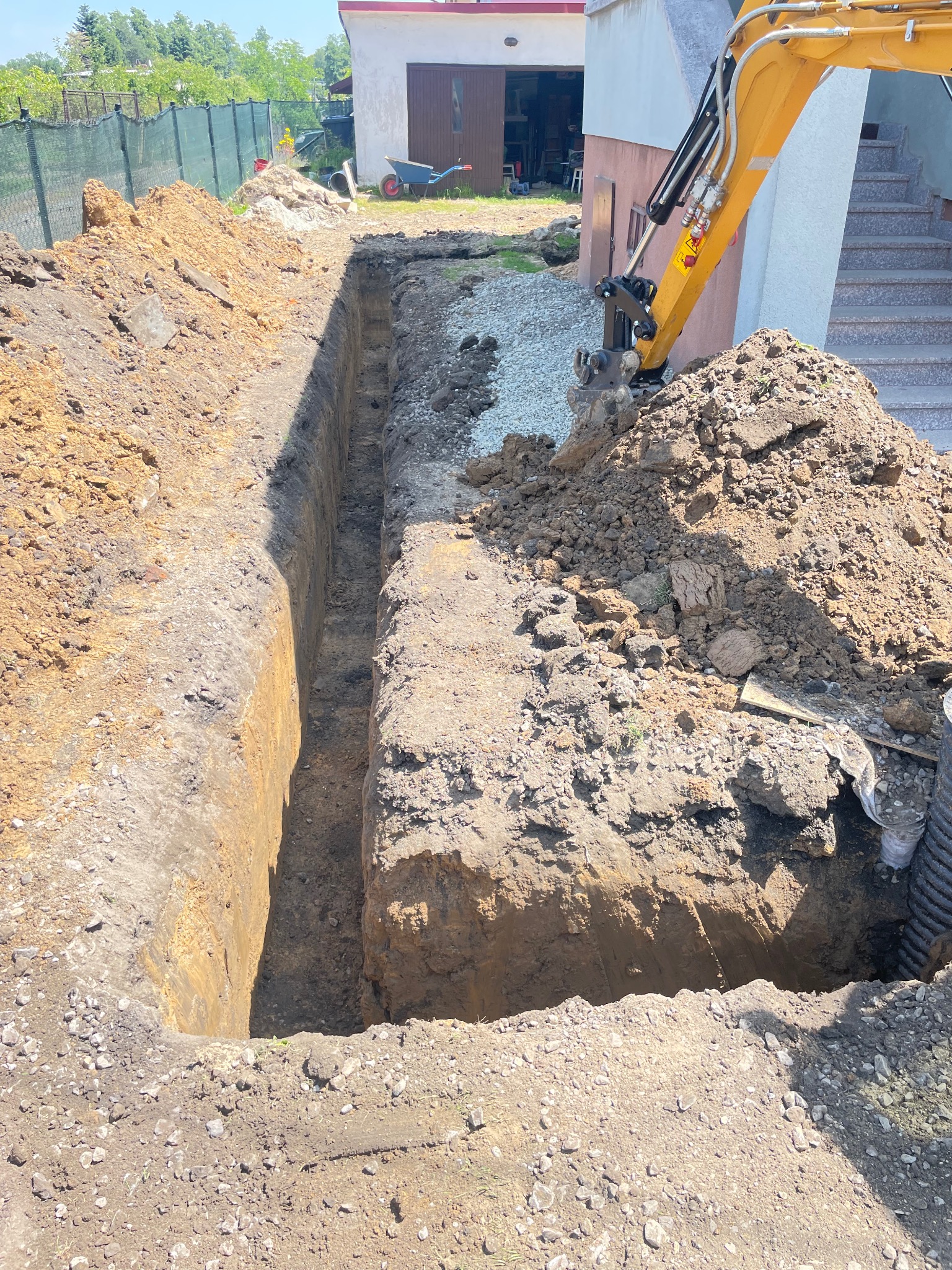Długi, wąski wykop w ziemi, obok budynku z żółtą koparką. Widoczna sterta ziemi i gruzu, w tle ogrodzenie i budynek gospodarczy.