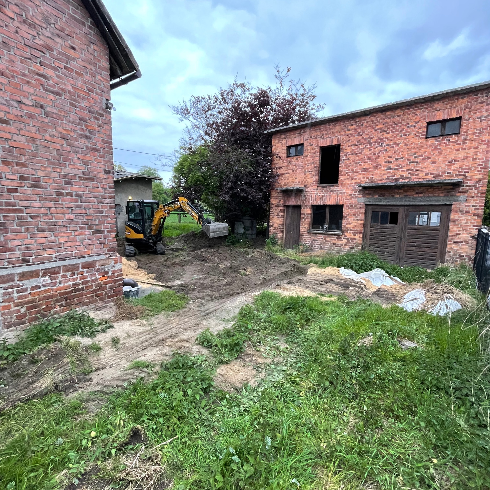 Żółta minikoparka w trakcie prac ziemnych przy ceglanym budynku, widoczne ślady opon na ziemi i częściowo usunięta trawa. Prace przygotowawcze pod budowę.