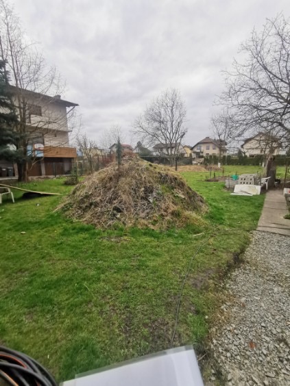 Kupa ziemi i roślin w ogrodzie, częściowo na trawie, z widocznymi budynkami w tle. Pochmurne niebo. Kable w lewym dolnym rogu kadru.